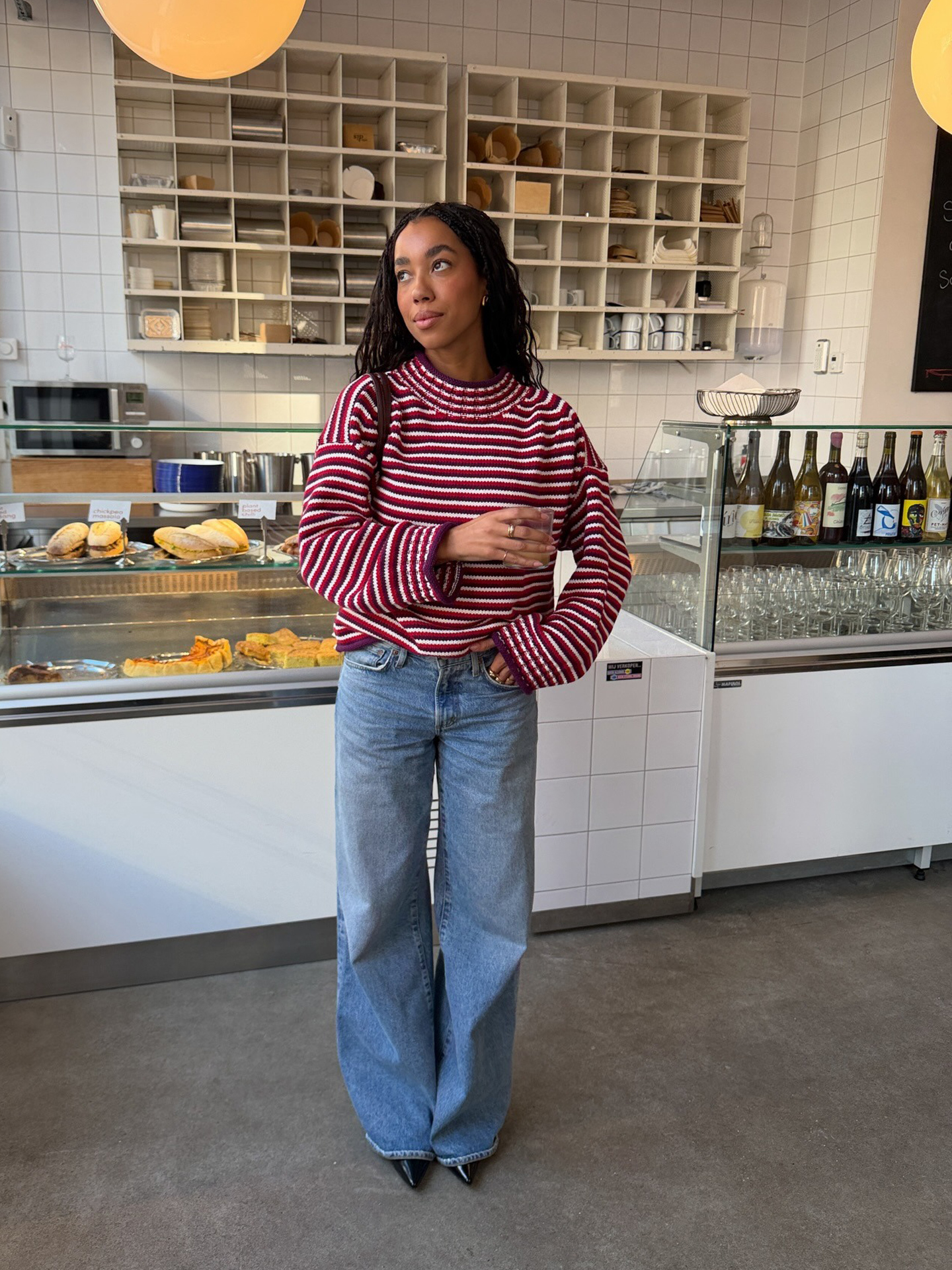 Dutch style influencer Amaka Hamelijnck poses in an Amsterdam coffee shop wearing a striped red J.Crew sweater, low-rise baggy jeans, and pointed black shoes