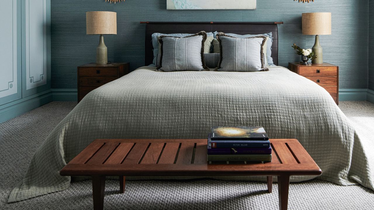 Blue-drenched bedroom with gray wall-to-wall carpeting. There are two wooden night stands with decorative table lamps, and a wooden bench in front of the bed.