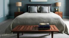 Blue-drenched bedroom with gray wall-to-wall carpeting. There are two wooden night stands with decorative table lamps, and a wooden bench in front of the bed.