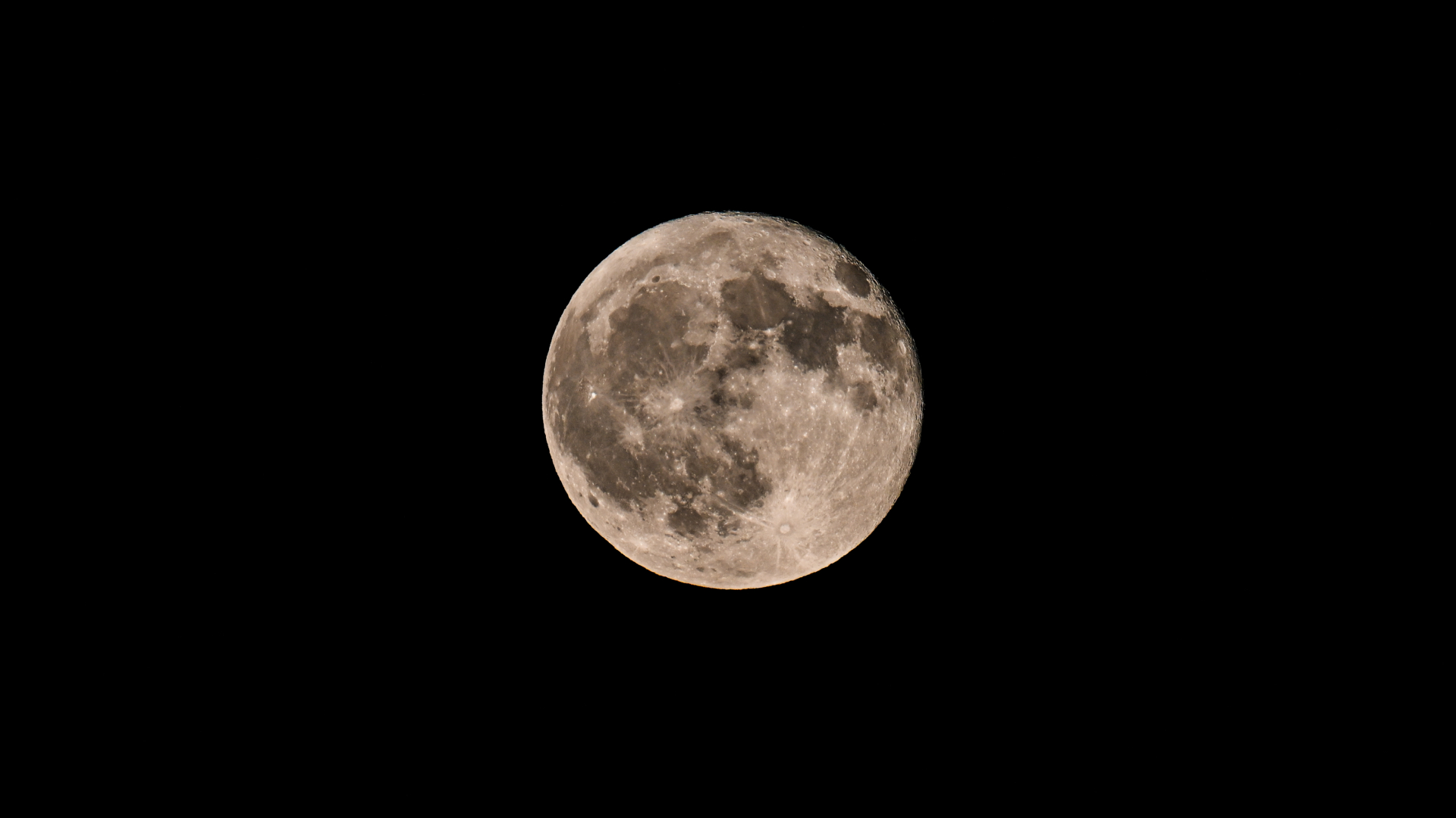 A detailed shot of a full moon hanging against a black night sky. Swathes of the lunar disk are covered in dark basaltic plains known as lunar seas. Impact craters are also visible marking the lunar surface.