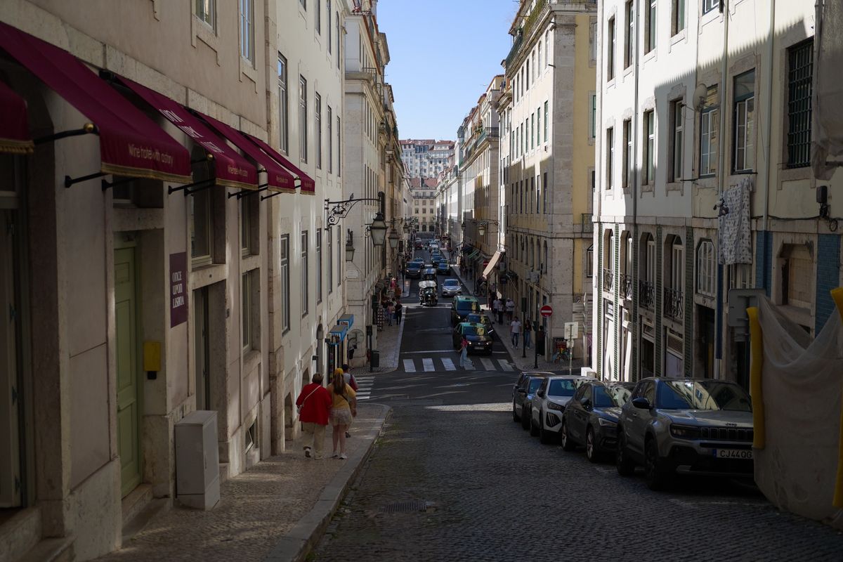 View down a narrow, sloping Lisbon street lined with historic buildings, parked cars and pedestrians.
