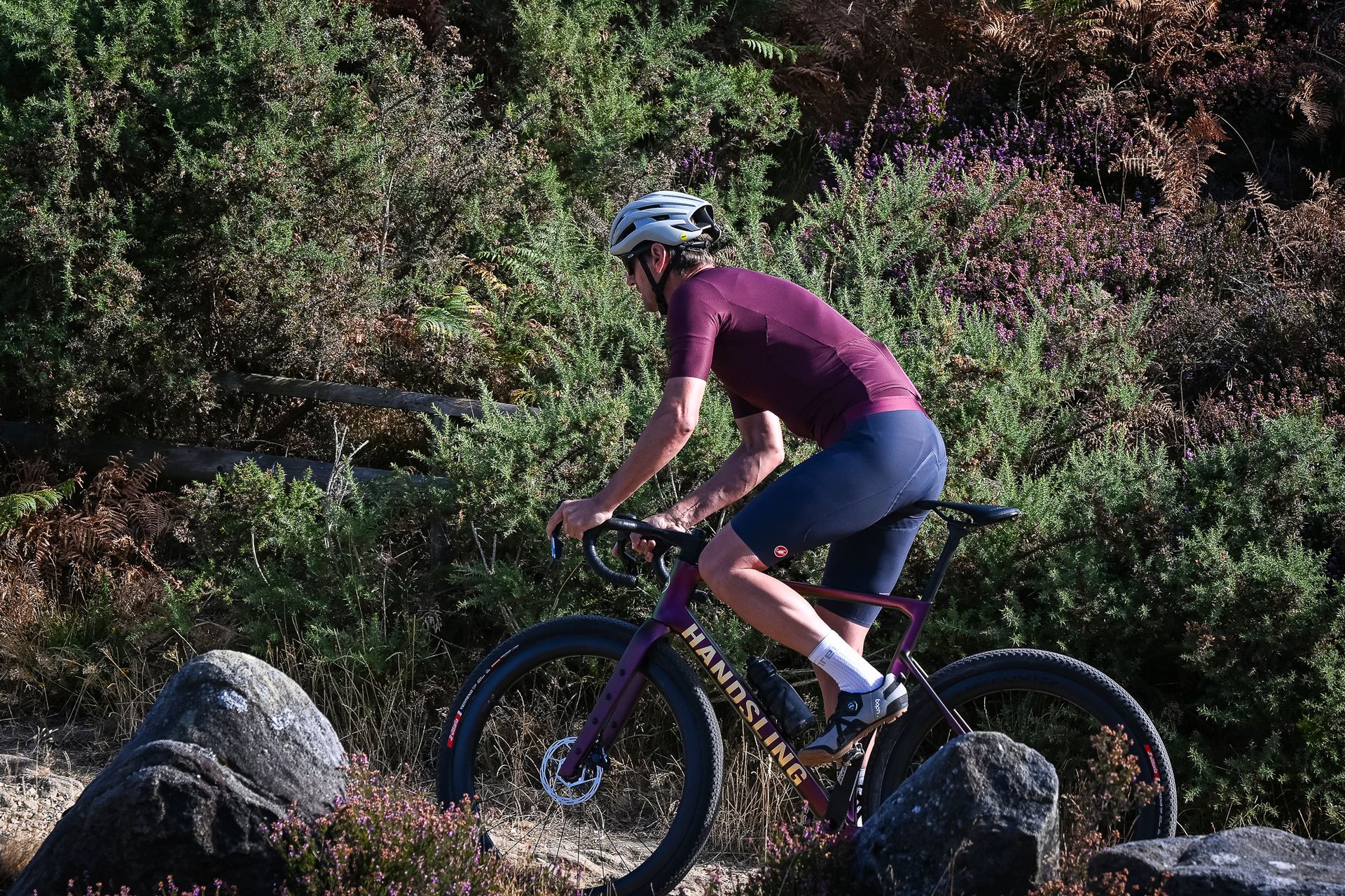Man riding a purple gravel bike up a rocky wearing grey shoes, blue shorts, a purple top and a beige helmet.