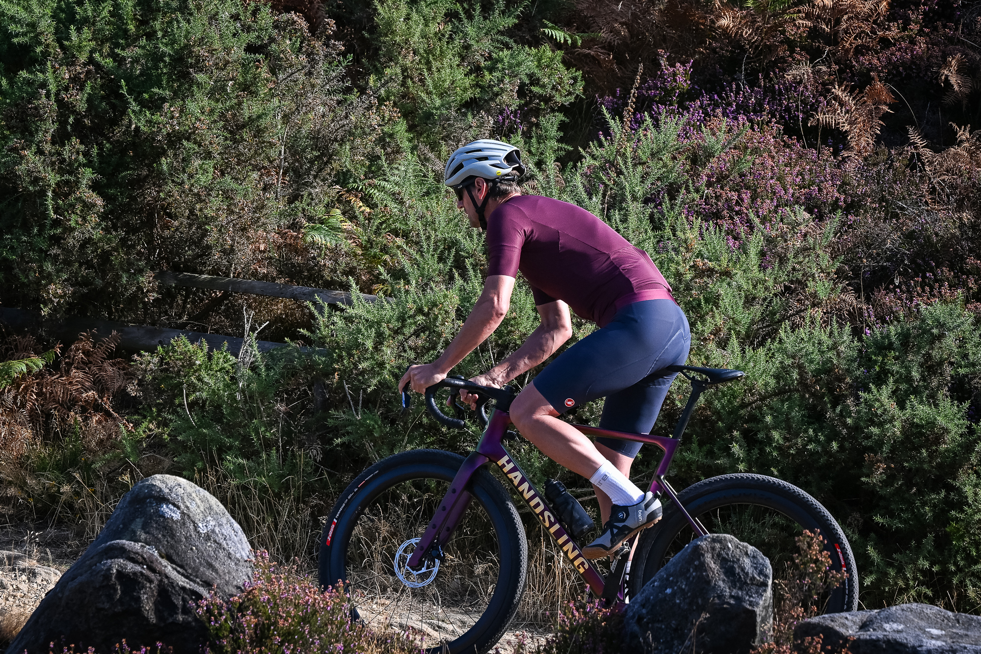 Man riding a purple gravel bike up a rocky wearing grey shoes, blue shorts, a purple top and a beige helmet.