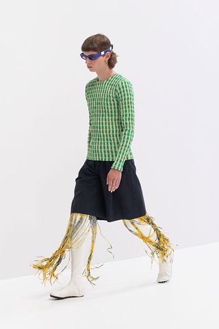A model walking down a white runway wearing a green and white checkered top, a black knee high skirt and white boots with yellow tassels on them.