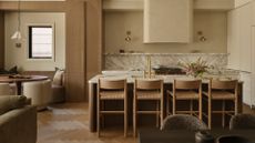 A beautiful kitchen with a marble backsplash, a stone island, a set of four beige woven chairs, brass sconces, a breakfast nook, and a scalloped pendant