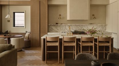 A beautiful kitchen with a marble backsplash, a stone island, a set of four beige woven chairs, brass sconces, a breakfast nook, and a scalloped pendant