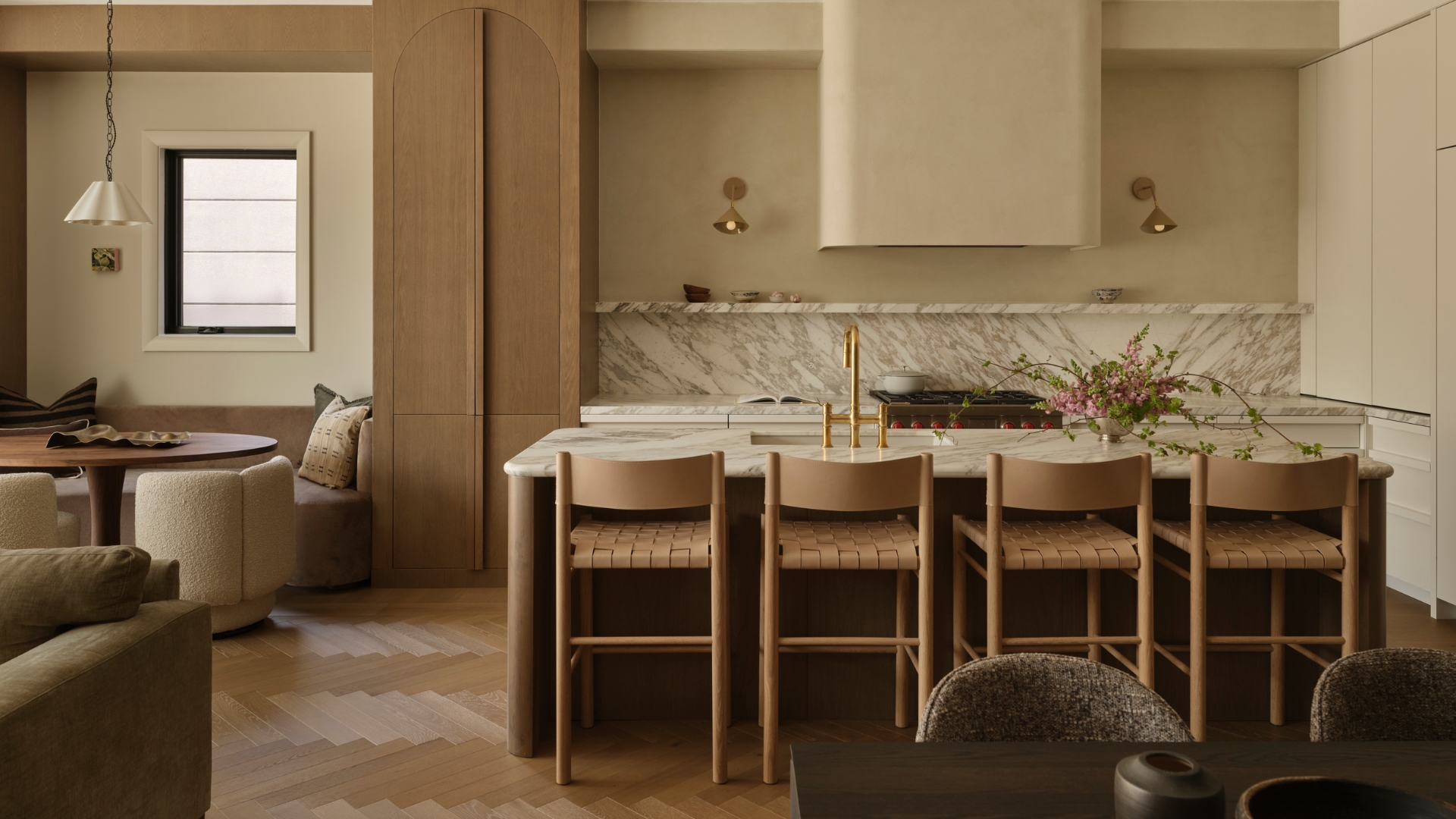 A beautiful kitchen with a marble backsplash, a stone island, a set of four beige woven chairs, brass sconces, a breakfast nook, and a scalloped pendant