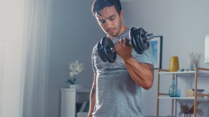 Strong Athletic Fit Man in T-shirt and Shorts is Doing bicep curls Exercises with Dumbbells at Home in His Spacious and Bright Apartment with Minimalistic Interior