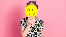An older woman holds a smiley face emoji in front of her face.