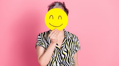 An older woman holds a smiley face emoji in front of her face.