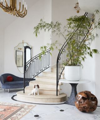 An entryway with curved staircase, black staircase railings and blue velvet chaise lounge with red velvet cushion