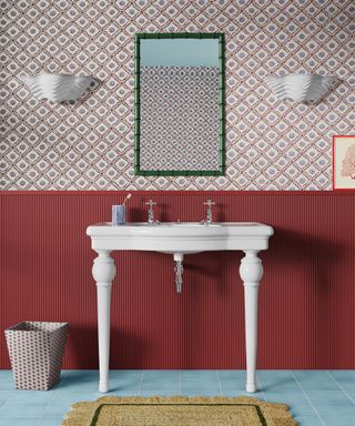 Colorful bathroom with floral wallpaper, red paneling, traditional white sink, blue square floor tiles, textured scalloped rug, two shell-style white wall lights, rectangular mirror with green frame.