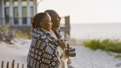 An older couple smile as they stand on the beach and share a blanket while drinking coffee, their beach house in the background.