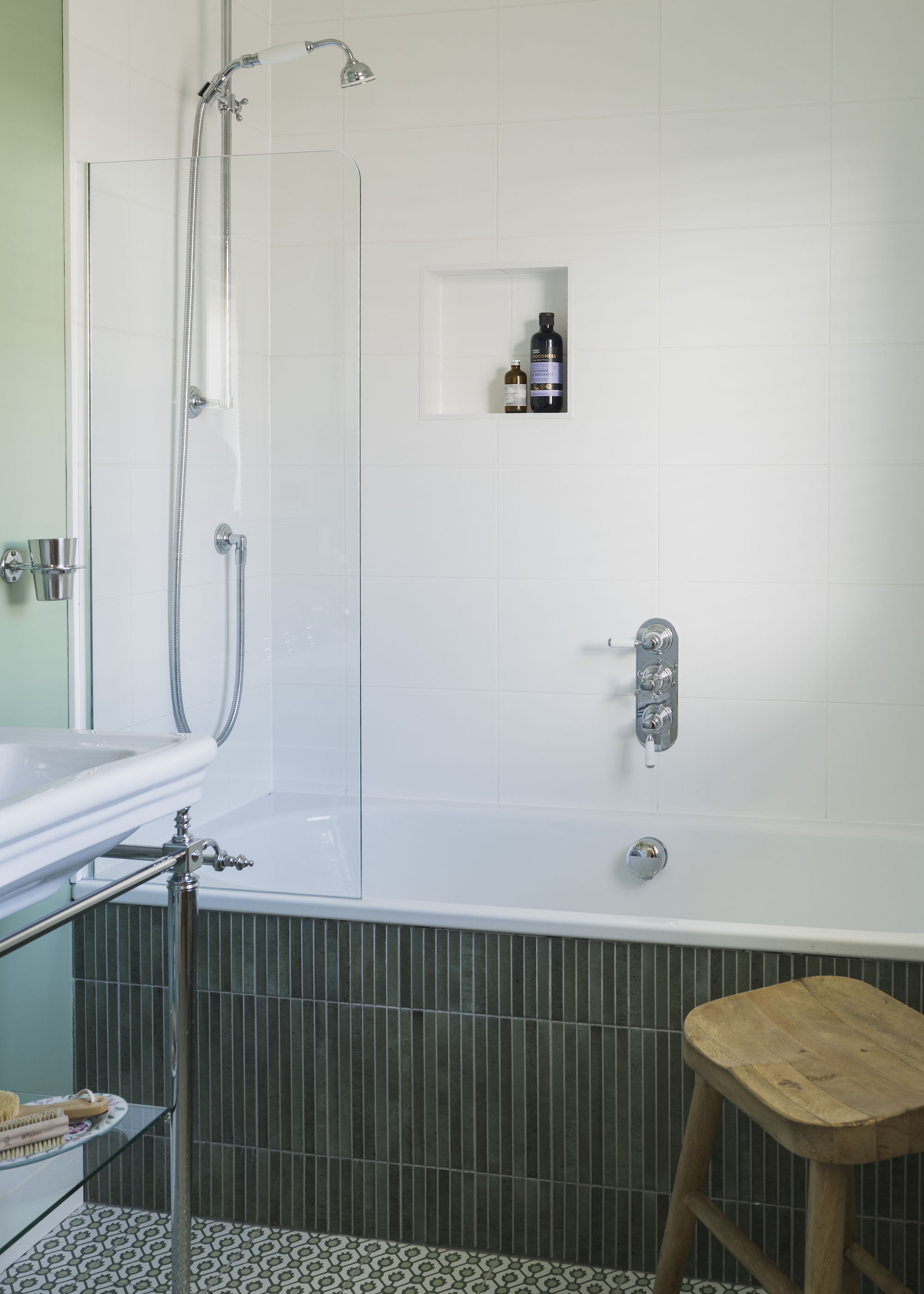 A bathtub covered in green tiles. Beside the bathtub is a wooden stool and a sink
