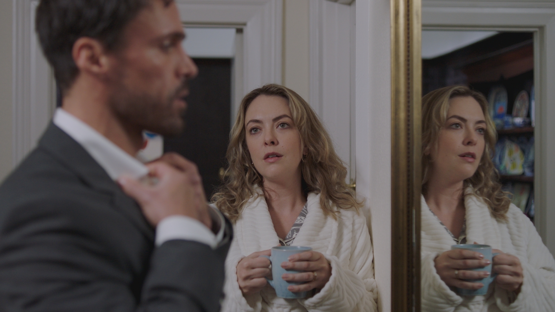 Flynn looking in the mirror wearing a suit, while his wife Claire looks on in her dressing gown.
