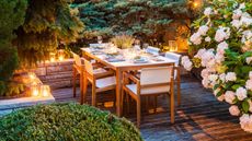 Patio with wooden dining table set for hosting dinner, it's surrounded by greenery and lit by glowing candles