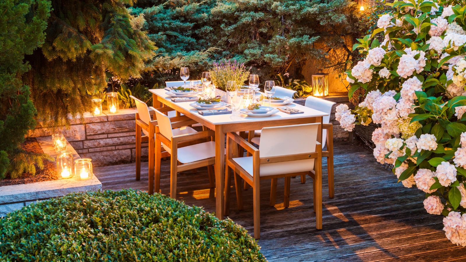 Patio with wooden dining table set for hosting dinner, it's surrounded by greenery and lit by glowing candles
