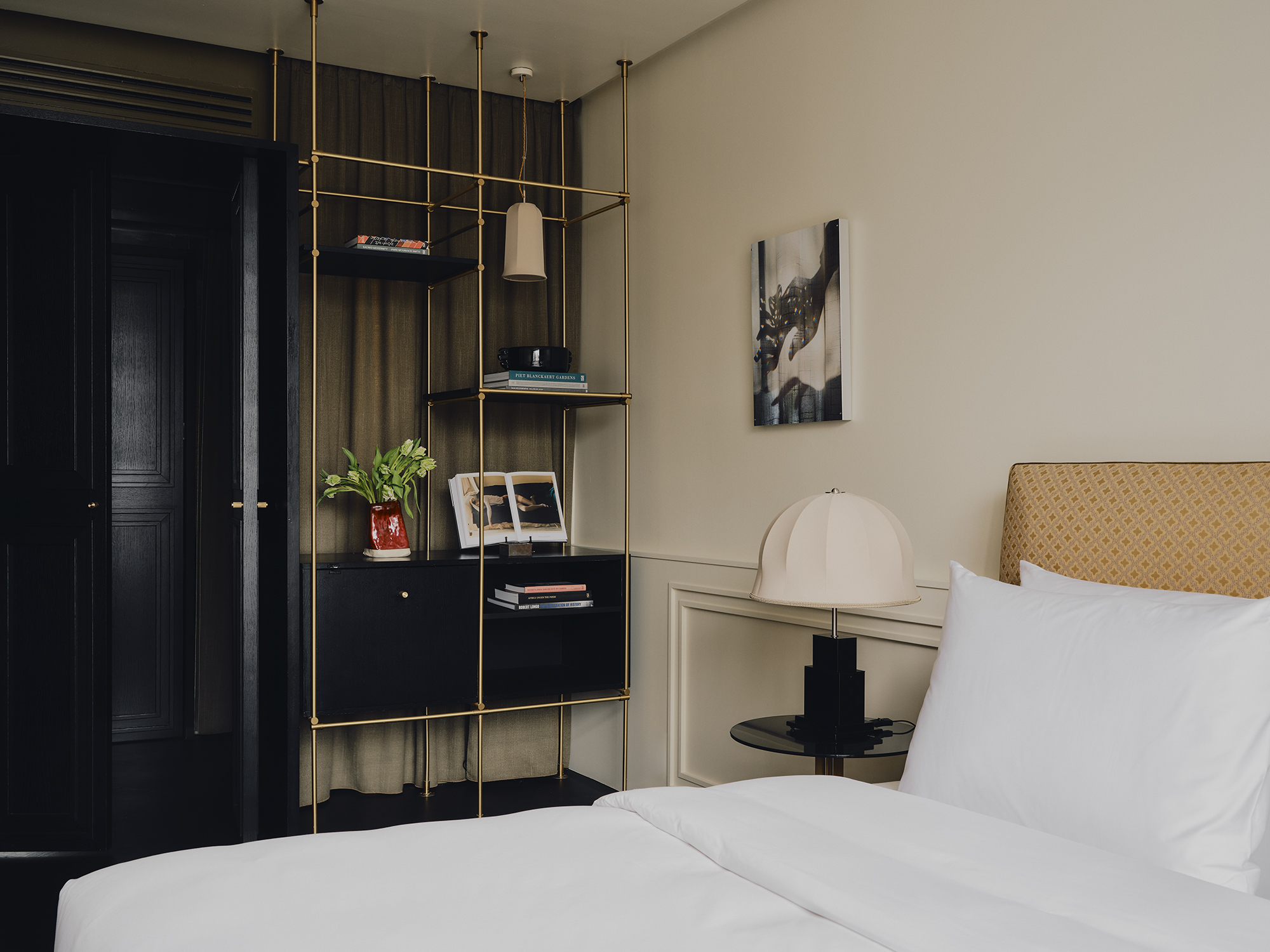 A hotel bedroom with white sheets and a butter yellow headboard with a modern table lamp on the bedside with a linen shade, next to open gold shelving in front of green curtains next to black doors