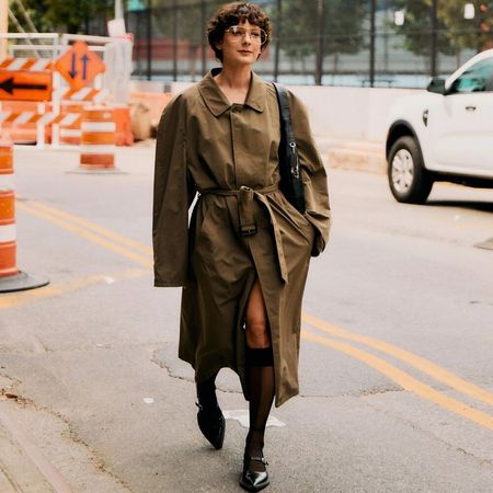 Alyssa wears a olive trench coat, sheer knee high socks, black flats, and a black handbag while walking the streets of NYC.