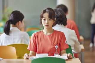 A woman wearing hospital scrubs (Park Bo-young as Jung Da-eun) sits at a cafeteria table with a food tray in front of her, in 'Daily Dose of Sunshine.'