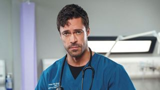 Olly as Casualty's new Clinical Lead Flynn Byron wearing blue scrubs and standing in a hospital, looking into the camera with a serious look on his face