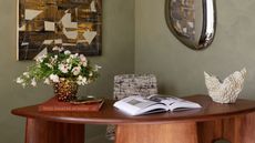 a green study with limewash walls, timber desk, artwork, vase, and open book on the desk