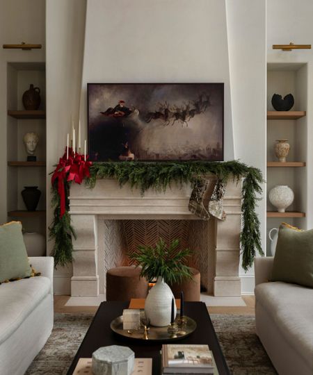 A cozy living room with a stone fireplace mantle draped with evergreen garland and a framed painting of Santa Claus and reindeer