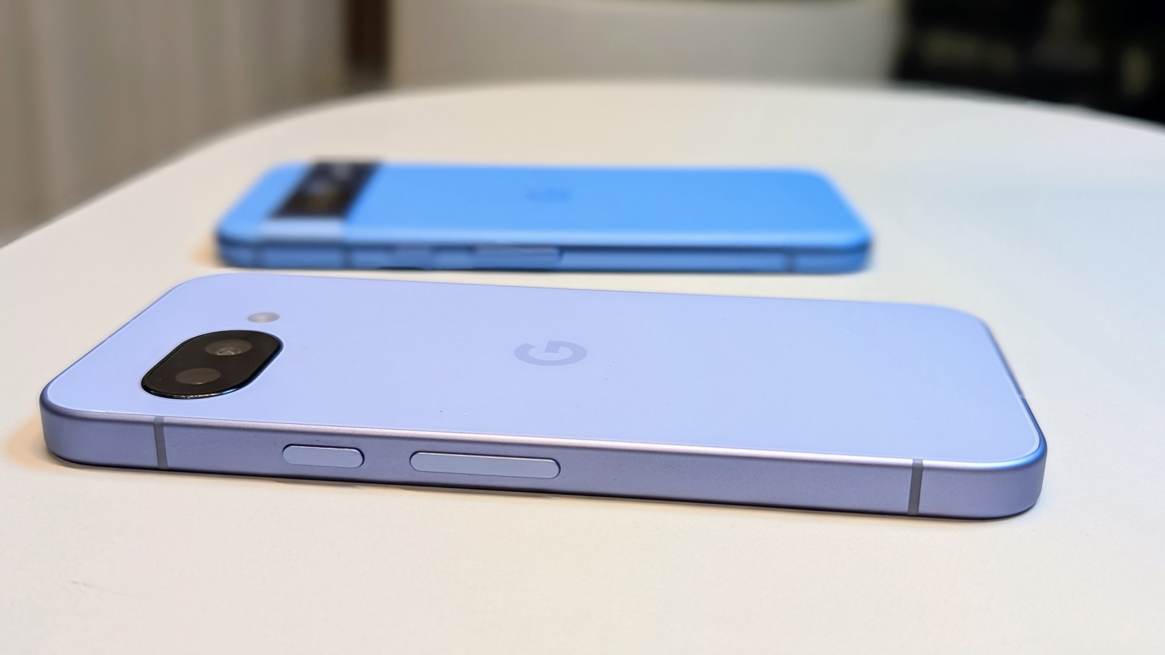 The Iris Google Pixel 9a (front) and Bay Google Pixel 8a (back) on a desk from a flat, close-up angle, showing the hard and soft edges of the 9a and 8a, respectively, and similar power/ volume rocker buttons.