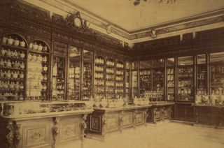 An olden day photo of a store with decorated wooden and glass counters in front of glass cabinets with products on rows of shelves inside them.