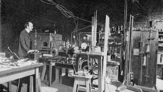 British physicist Joseph John Thomson standing in a laboratory filled with equipment.