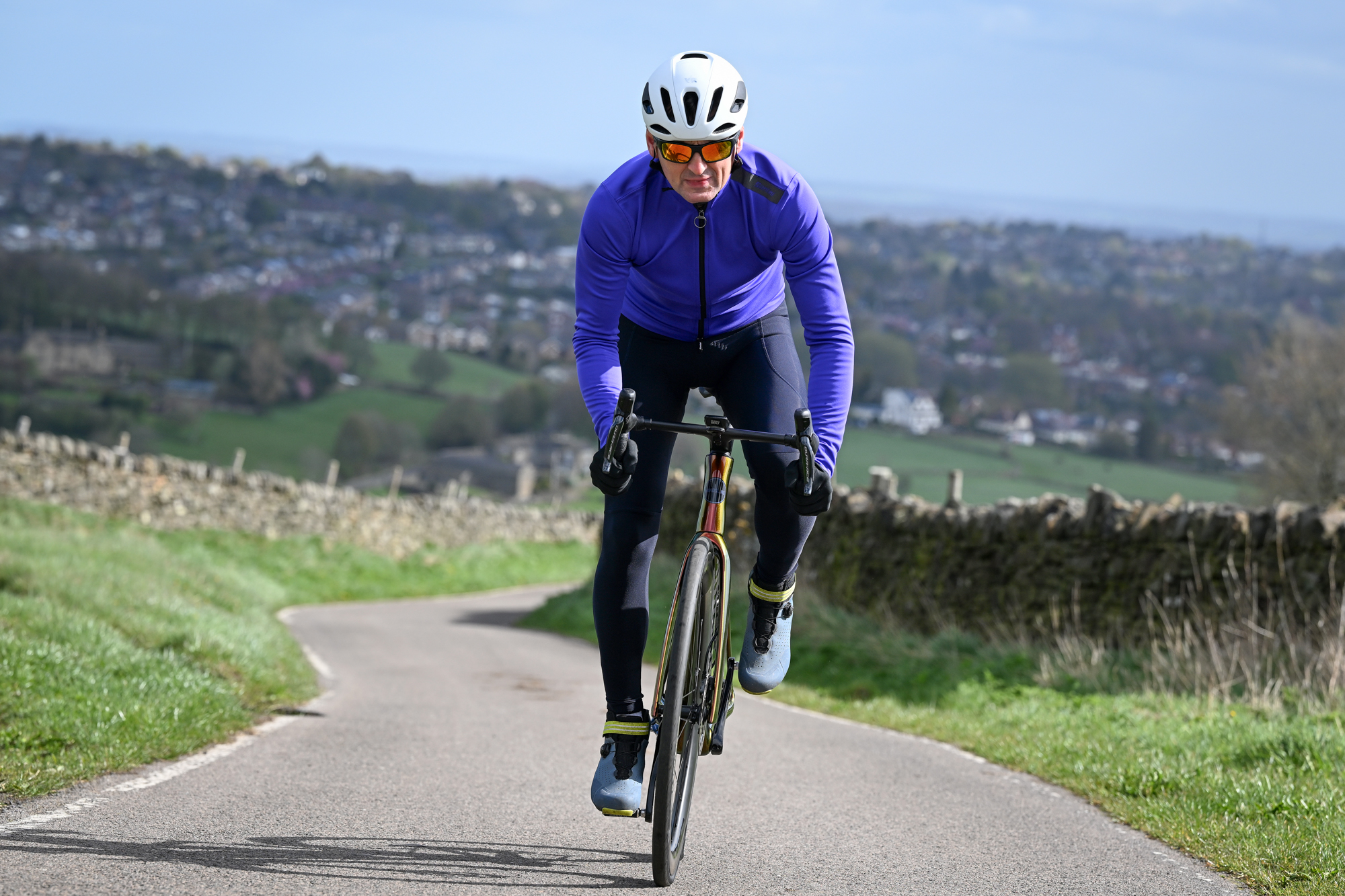 Man in a purple jacket, sunglasses, blue shoes and white helmet riding a bike up a country lane