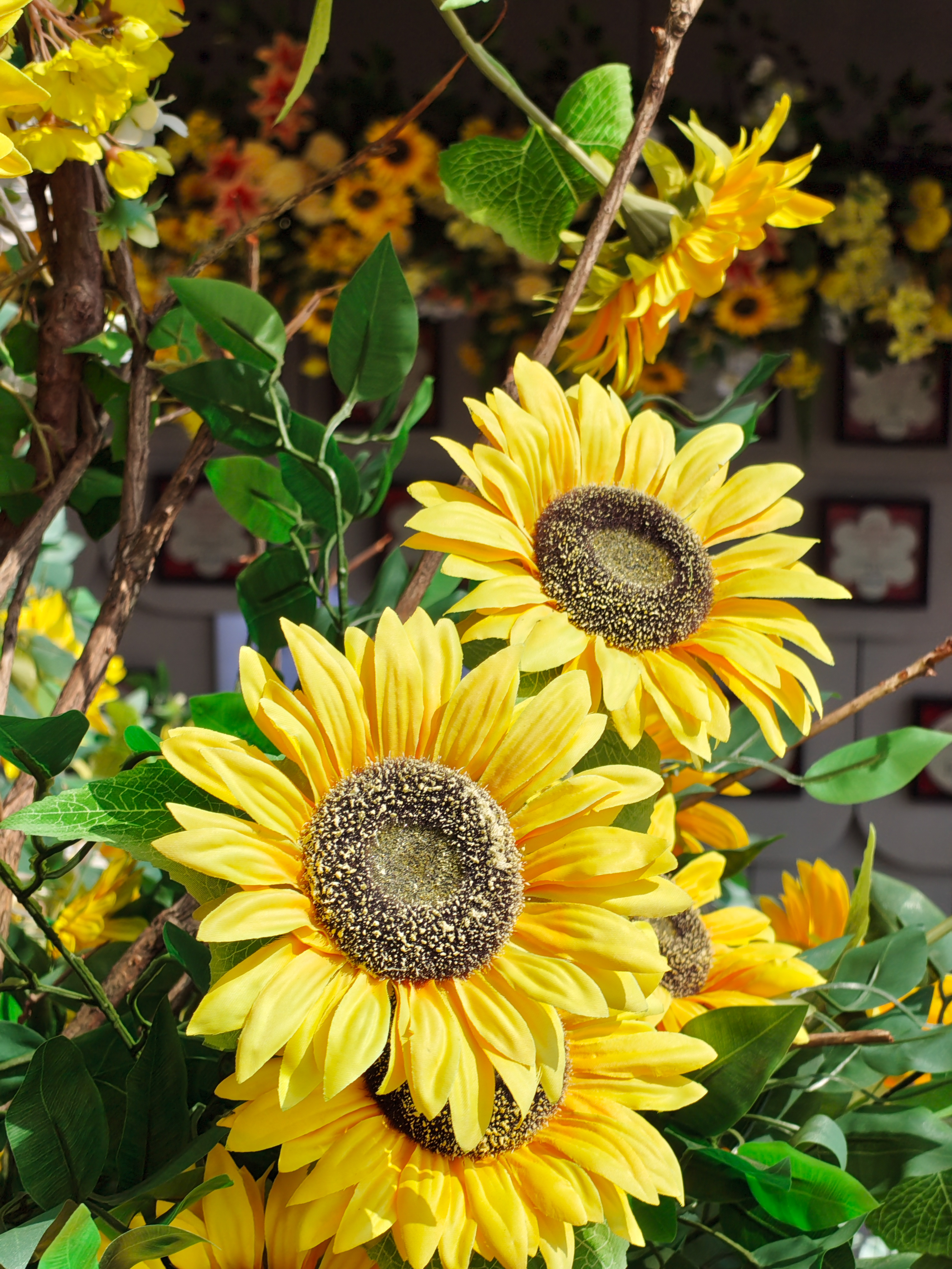 A vertical, close-up photograph of several bright yellow artificial sunflowers. The focus is sharp on two large sunflowers in the foreground, showing the detailed texture of their dark brown centers and layered yellow petals. Green leaves and thin brown branches are interspersed among the flowers, with more blurred sunflowers visible in the background.