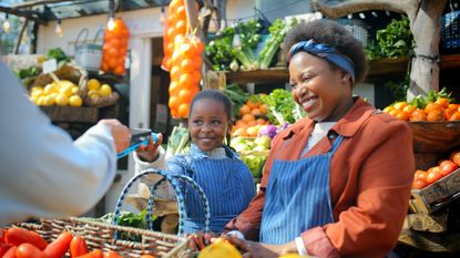 A business owner and her child smile while conducting a transaction with a farmers market customer.