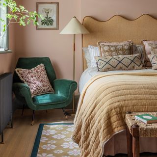 A dusky pink-painted bedroom with a beige headboard and matching bedcover and a green velvet armchair in the corner