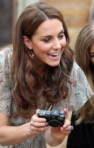 Kate Middleton wearing a paisley short sleeve dress leaning forward and holding a camera while smiling