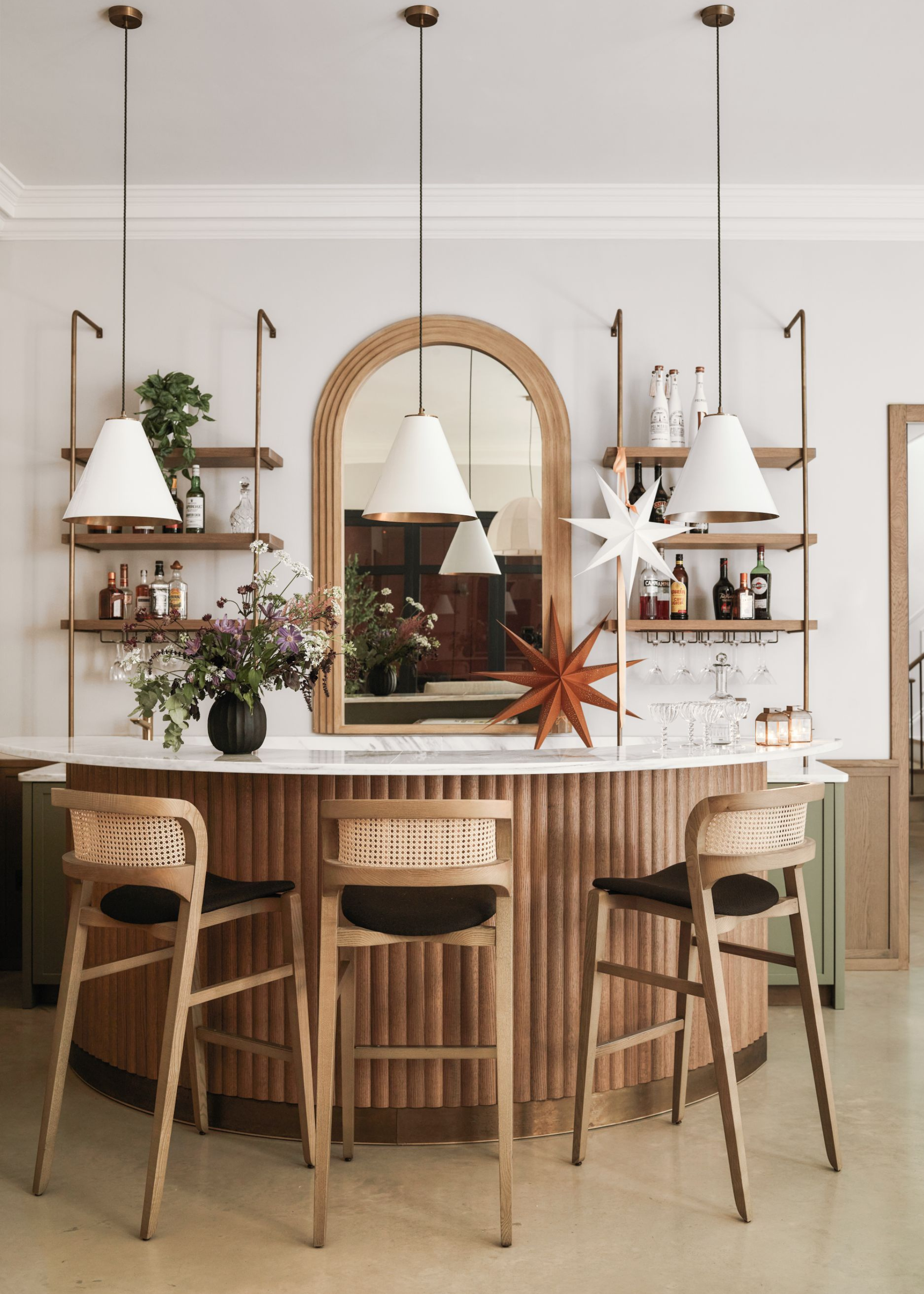 A stylish home bar with fluted wood island and a row of three white pendant lights above the counter; it has two paper star shades displayed behind and above the counter, one in orange and one in white