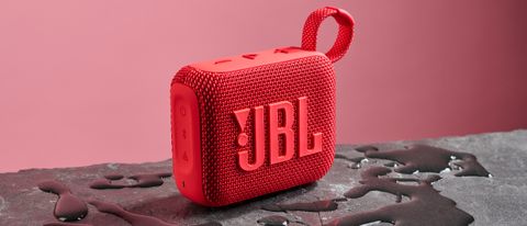 A red JBL Go 4 pictured against a pink background. It is sitting on a dark stone surface with puddles of water around it.