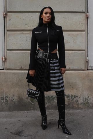 a photo showing what to wear to a rap concert with a woman wearing a cropped jacket with a plaid midi skirt and knee-high boots and a black shoulder bag