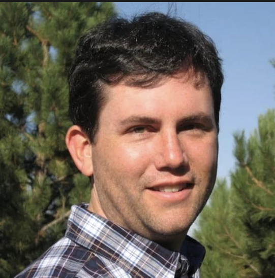Headshot of a man with short, curly dark brown hair wearing a check shirt