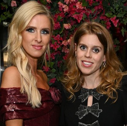 Nicky Hilton standing in front of a pink floral display wearing a burgundy sequined dress standing next to Princess Beatrice in a black dress covered in silver bows 