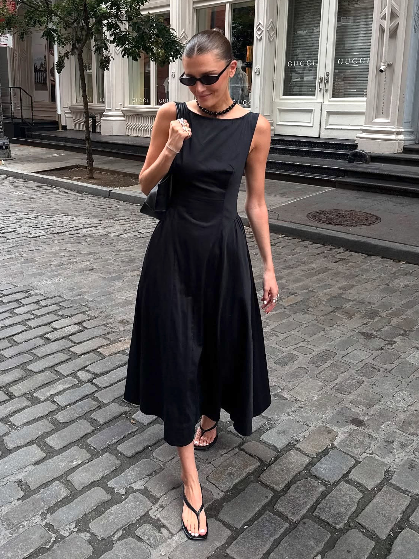 NYC style influencer Coco Schiffer walking on a cobblestone street wearing a black sleeveless a-line dress and black flip-flop sandals