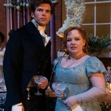 Luke Newton as Colin Bridgerton and Nicola Coughlan as Penelope Featherington, standing by a ball's drink table with another woman behind them