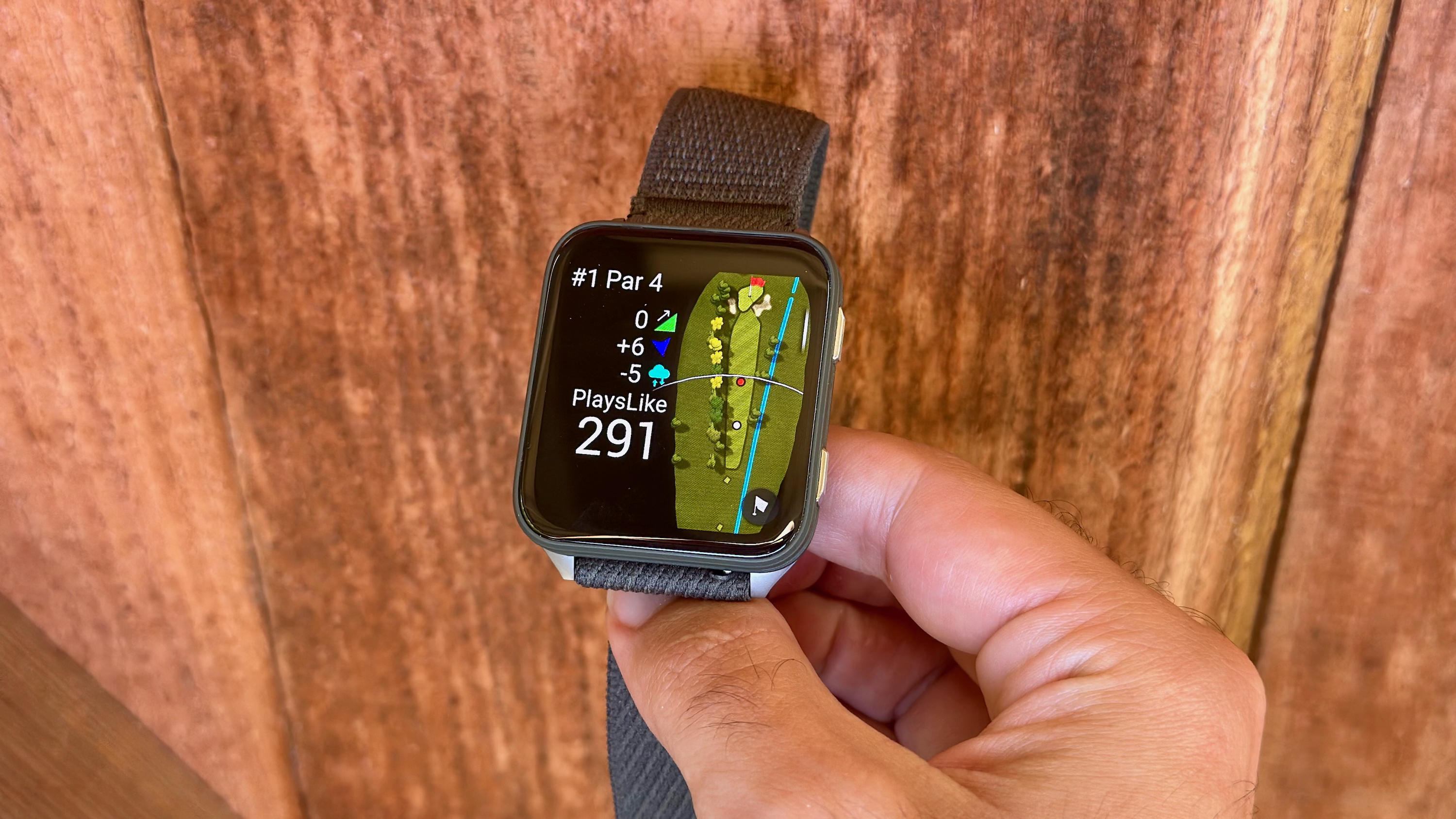 The Garmin Venu X1 showing a Golf activity with the Playslike distance visible, including elevation, wind, and humidity data. Behind the watch is a fence.