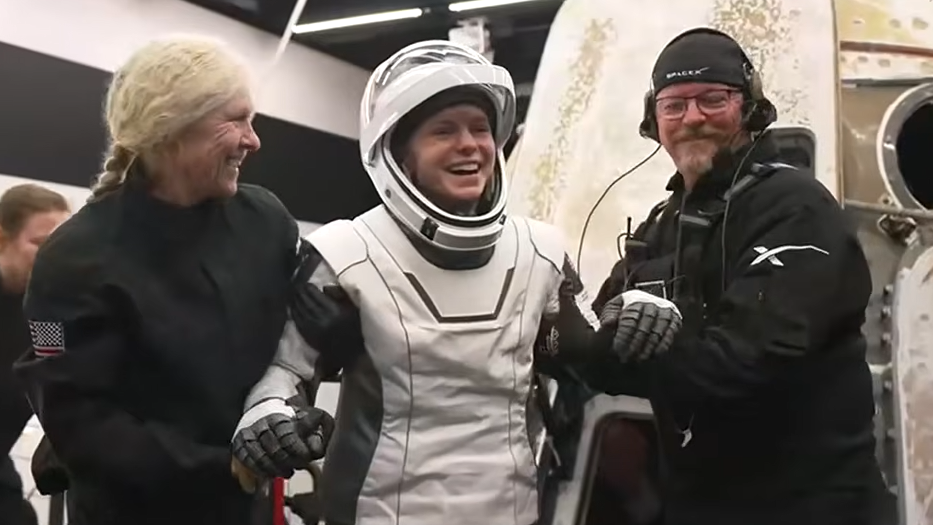 a woman in a white spacesuit smiles as she is helped to her feet