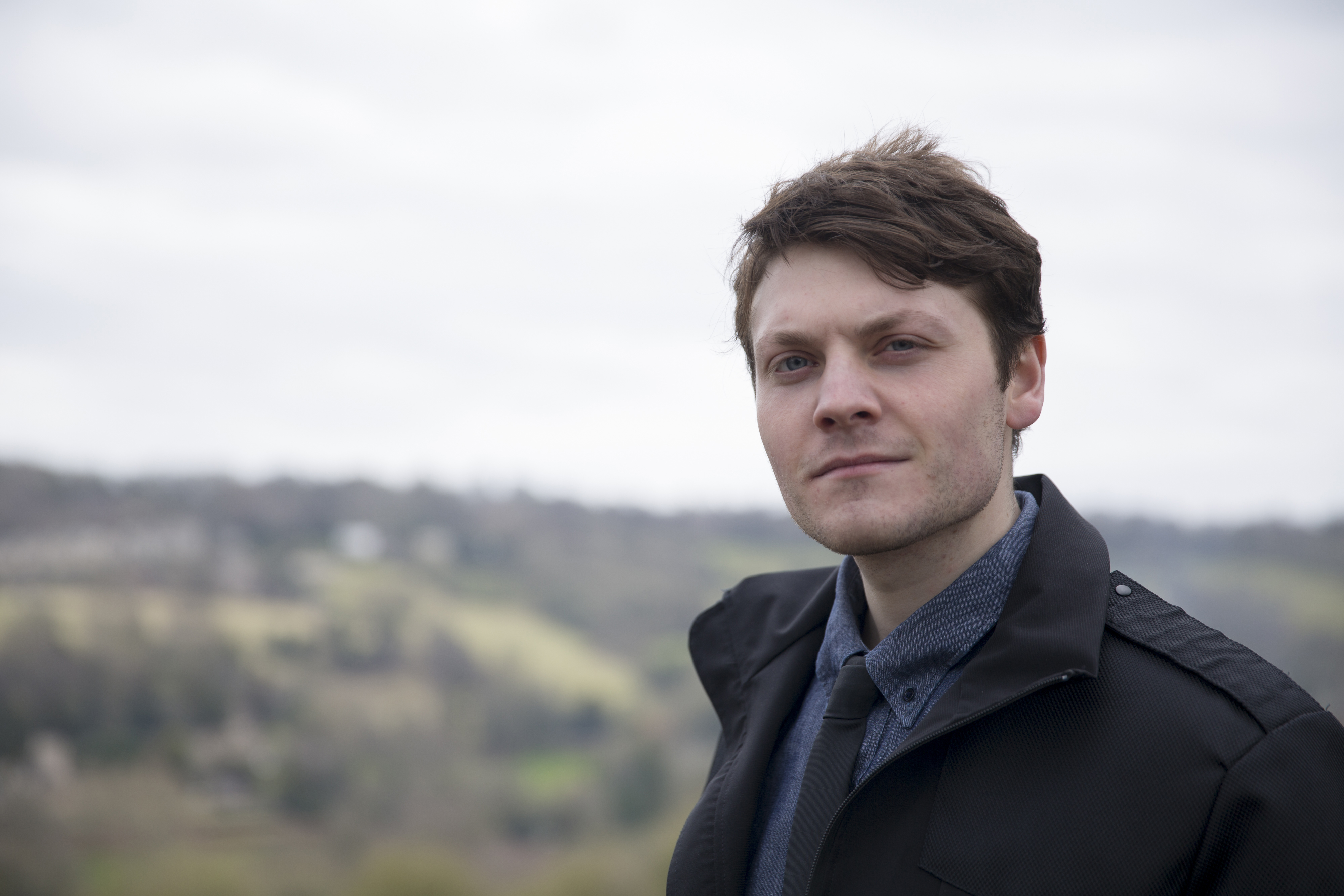 White man with dark hair, shirt and tie and black jacket, posing against a backdrop of a hillside and sky