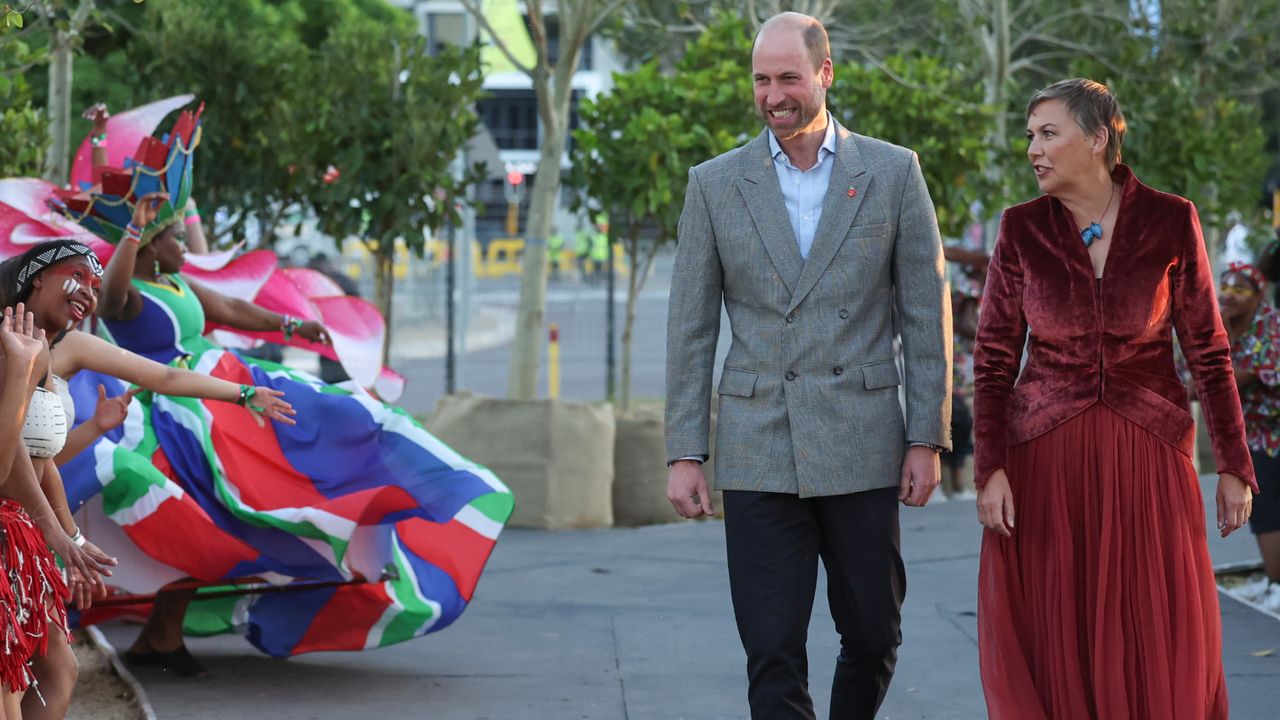 Prince William wearing a gray blazer walking next to the Earthshot Prize Chief Executive Hannah Jones in a burgundy dress watching local dancers in Cape Town, South Africa