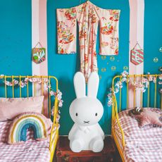 A blue-painted shared children's room with two yellow metal beds and a Miffy light and a kimono hung on the wall in the middle