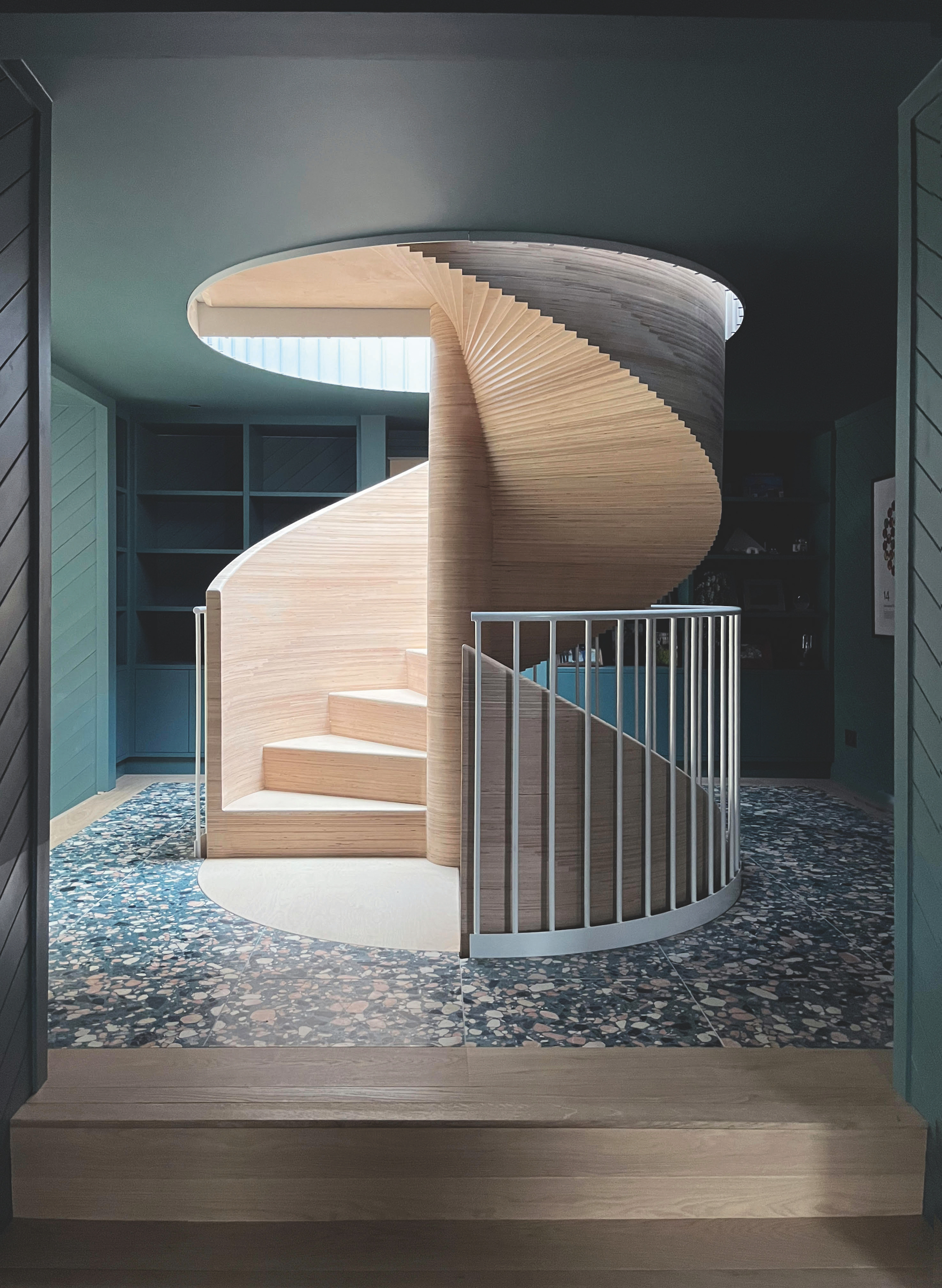 A light-wood spiral staircase descending from a circular hole in the ceiling and into the floor below, with a white railing around.