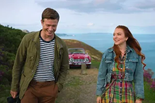a man (ed speelers) and a woman (lindsay lohan) stand in a grassy field in front of a red classic car
