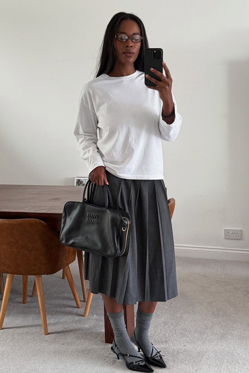 a woman wearing a white longsleeve, gray pleated skirt, gray socks, miu miu back bag, and black heels